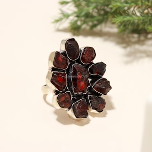 May include: A silver ring featuring a cluster of raw, deep red garnet stones. The gemstones are set in individual silver bezels, forming a floral or starburst design. The ring has a wide band and a polished finish.