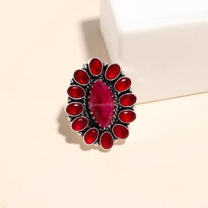 Könnte beinhalten: Ein silberner Ring mit einem großen, oval geformten, tiefroten Edelstein in der Mitte, umgeben von einem Kranz aus kleineren, ovalen roten Steinen. Der Ring hat ein detailliertes, strukturiertes Silberband.