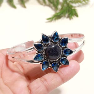 May include: Silver cuff bracelet with a blue gemstone center surrounded by smaller blue gemstones in a flower shape.