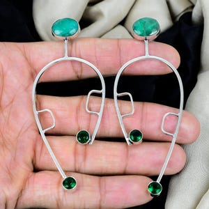 May include: A pair of silver earrings with a distinctive design. Each earring has a large oval green gemstone at the top, a curved silver frame, and three smaller green gemstones. The earrings are held in a hand.