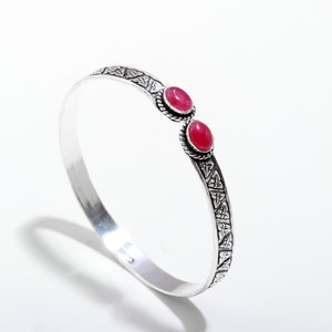 May include: Silver-toned cuff bracelet with two oval, red gemstones. The bracelet features a detailed Celtic knot design on the ends and around the gemstones. The bracelet is a stylish piece of jewelry.