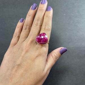 May include: A gold-toned ring featuring a large, faceted, heart-shaped ruby gemstone. The ruby displays a deep red hue with lighter pink facets. The ring is set on a hand with purple nail polish.
