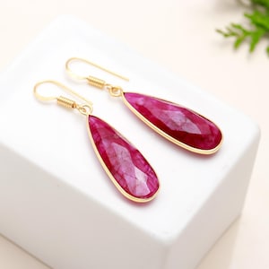 Op de afbeelding: Een paar goudkleurige oorhangers met druppelvormige, gefacetteerde, robijnrode edelstenen. De oorbellen hebben een goudkleurig montuur rond de edelstenen en goudkleurige oorhaken met een spiraalvormig ontwerp.