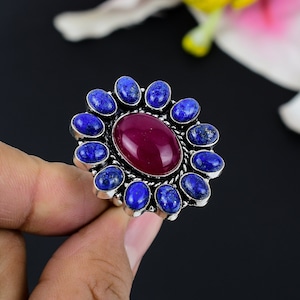 May include: A silver ring featuring a large oval, deep red gemstone at the center, surrounded by a halo of smaller, oval, blue gemstones. The ring is set against a dark background, highlighting the vibrant colors of the stones.