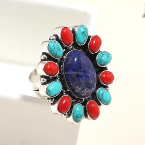 May include: A silver ring featuring a large oval blue stone at the center, surrounded by alternating red and turquoise stones. The ring has a detailed, ornate design, with the text "@JewelryByDVS" visible.
