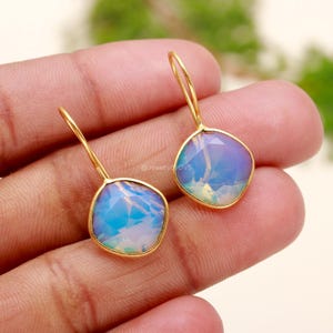 May include: A pair of gold drop earrings featuring iridescent blue and white opal stones. The stones are set in a gold bezel setting and the earrings have a simple, elegant design.