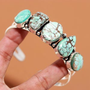May include: A silver cuff bracelet featuring five polished, light green turquoise stones with dark brown matrix patterns. The stones are set in smooth silver bezels and separated by small silver spheres along the band of the jewellery piece.