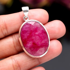 May include: Oval-shaped pendant with a large, faceted, deep pink gemstone set in a silver-colored metal frame. The pendant has a simple bail for attaching to a chain. The gemstone's surface reflects light, showcasing its cut.