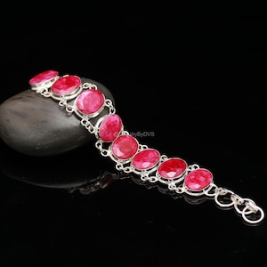 Peut inclure: Un bracelet en argent orné de pierres précieuses rouges ovales. Les pierres sont serties dans un design en chaîne, créant un accessoire audacieux et élégant. Le bracelet est présenté sur une surface sombre et réfléchissante.