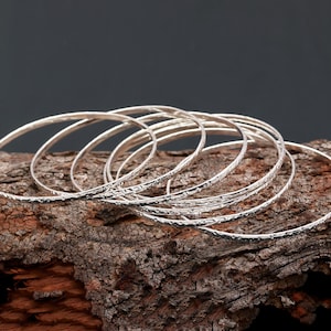 Può includere: Un set di braccialetti d'argento con una superficie testurizzata. I braccialetti sono impilati uno sopra l'altro e disposti in un cerchio.