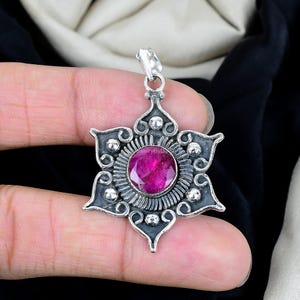 May include: A silver pendant with a starburst design, featuring a large, round, deep pink gemstone at its center. The pendant has intricate detailing and a bail for a chain. The gemstone is surrounded by a textured silver setting.