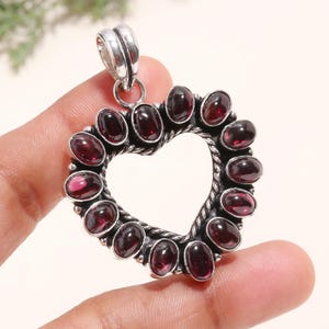 Puede incluir: Colgante en forma de corazón con marco de metal plateado. El colgante está adornado con piedras preciosas ovaladas de color rojo intenso. Las piedras están dispuestas alrededor del contorno del corazón, creando contraste con el metal.