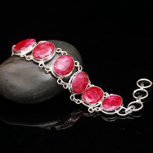 Può includere: Un bracciale in argento con pietre preziose rosse ovali e vibranti. Le pietre sono incastonate in castoni individuali in argento e collegate tra loro. Il bracciale è esposto su una superficie scura e riflettente, evidenziando il colore e il design.