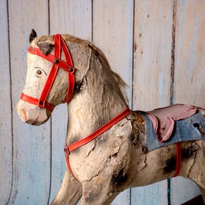 May include: A vintage wooden rocking horse with a red bridle and a blue and pink saddle. The horse is weathered and shows signs of age.