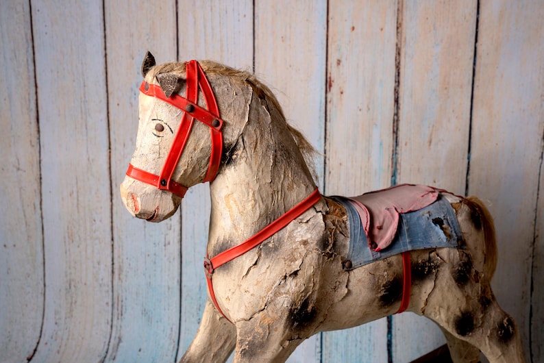May include: A vintage wooden rocking horse with a red bridle and a blue and pink saddle. The horse is weathered and shows signs of age.