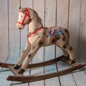 May include: A vintage wooden rocking horse with a worn white paint finish and red leather straps. The horse has a blue and pink saddle and a wooden base.