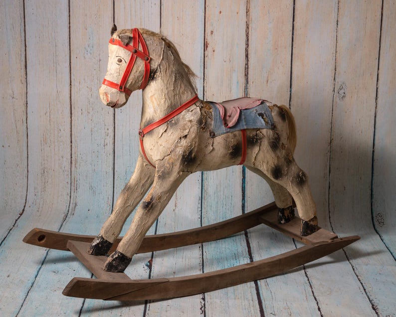 May include: A vintage wooden rocking horse with a worn white paint finish and red leather straps. The horse has a blue and pink saddle and a wooden base.