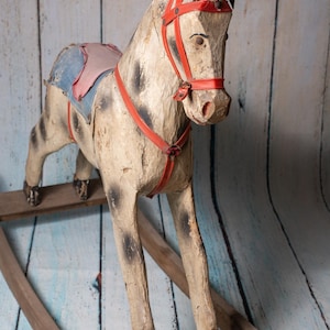 May include: A vintage wooden rocking horse with a red leather bridle and saddle. The horse is painted white with black spots and has a worn, rustic look.