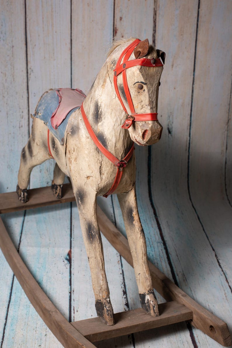 May include: A vintage wooden rocking horse with a red leather bridle and saddle. The horse is painted white with black spots and has a worn, rustic look.
