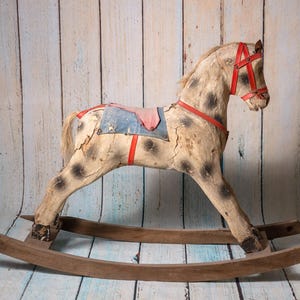 May include: A vintage wooden rocking horse with a worn white paint finish and red leather accents. The horse has a blue and red saddle and a brown wooden base.