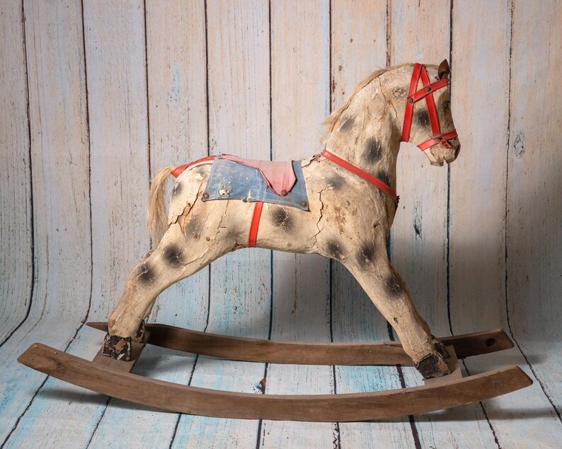 May include: A vintage wooden rocking horse with a worn white paint finish and red leather accents. The horse has a blue and red saddle and a brown wooden base.