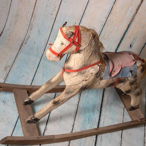 May include: A vintage wooden rocking horse with a red and white bridle and a blue and pink saddle. The horse is weathered and has a rustic look.