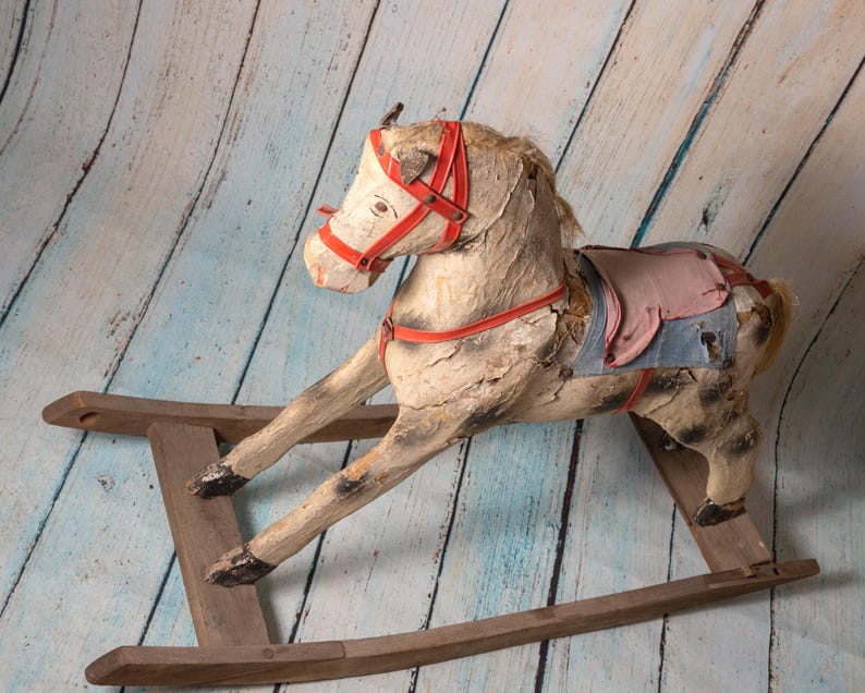May include: A vintage wooden rocking horse with a red and white bridle and a blue and pink saddle. The horse is weathered and has a rustic look.