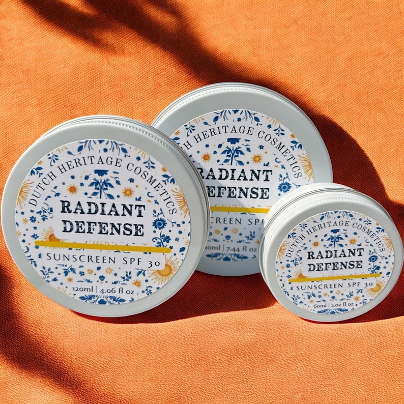 Op de afbeelding: Drie ronde, lichtblauwe metalen containers met Radiant Defense zonnebrandcr&egrave;me SPF 30 van Dutch Heritage Cosmetics. De containers hebben witte etiketten met blauwe bloemen- en zonnemotieven. De grootste container bevat 120 ml.