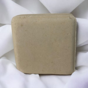 Puede incluir: Una barra de jab&oacute;n beige cuadrada sobre un fondo de tela blanca.