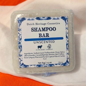 Puede incluir: Una barra de champ&uacute; blanca con una etiqueta azul y blanca que dice "Dutch Heritage Cosmetics Shampoo Bar Unscented". La etiqueta tambi&eacute;n incluye los ingredientes, el peso neto de 65 g (2,3 oz) y el texto "Animal Based Handmade".