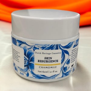 Op de afbeelding: Een witte pot met een blauw-witte bloemenlabel. Het label vermeldt "Dutch Heritage Cosmetics Skin Resurgence Chamomile Net 60ml | 2.1 fl oz".