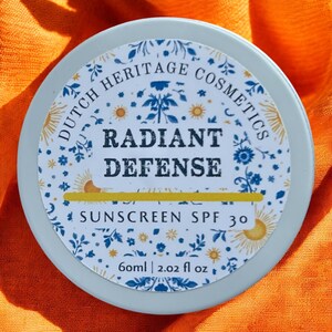 Op de afbeelding: Een ronde, lichtblauwe container van Dutch Heritage Cosmetics Radiant Defense Sunscreen SPF 30. Het etiket toont bloemen- en zonillustraties, met de tekst "Radiant Defense". De container bevat 60 ml.
