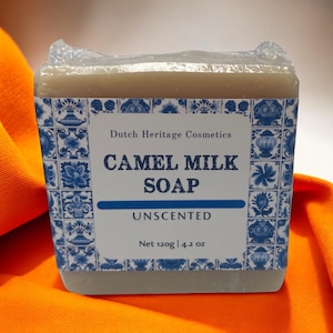 May include: A bar of unscented camel milk soap wrapped in plastic. The soap is beige and has a blue and white label with the text "Dutch Heritage Cosmetics Camel Milk Soap Unscented Net 120g | 4.2 oz".
