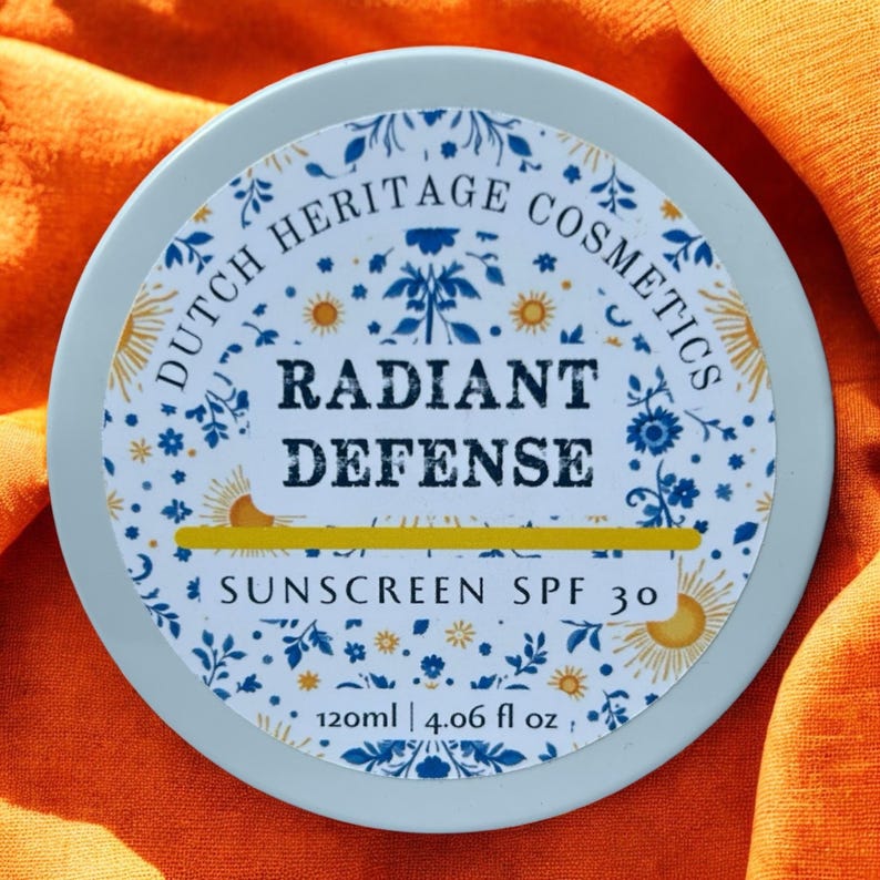 Op de afbeelding: Een ronde, lichtblauwe container met zonnebrandcr&egrave;me en een decoratief etiket. Het etiket bevat de tekst "DUTCH HERITAGE COSMETICS" en "RADIANT DEFENSE SUNSCREEN SPF 30". De container bevat 120 ml (4,06 fl oz).