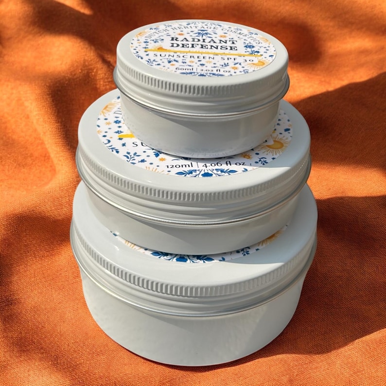 Op de afbeelding: Drie gestapelde, lichtgrijze metalen blikken met schroefdeksels. De deksels hebben een wit etiket met blauwe bloemmotieven en de tekst "Radiant Defense Sunscreen SPF 30". De blikken staan op een oranje stoffen achtergrond.