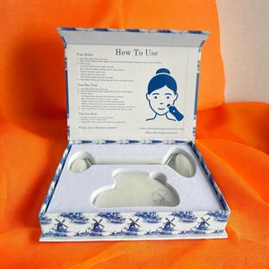 May include: A white box with a blue and white windmill pattern. The box contains a white jade roller and gua sha tool. The box lid has instructions on how to use the tools. The text on the lid reads "How To Use" and includes instructions for "Face Roller" and "Gua Sha Tool".