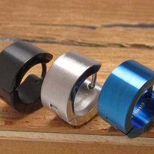 Pode incluir: Três brincos de argola em aço inoxidável preto, prateado e azul. Os brincos têm um acabamento em metal escovado e um design articulado. Os brincos são exibidos em uma superfície de madeira.