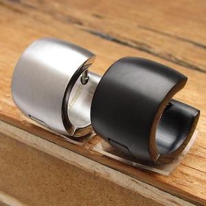 Può includere: Due anelli a polsino, uno in acciaio inossidabile e uno nero. Gli anelli hanno un design curvo e arrotondato. L'anello argentato è in acciaio inossidabile spazzolato, e l'altro è nero. Sono esposti su una superficie di legno.