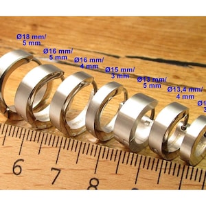 Pode incluir: Uma coleção de brincos de argola prateados de vários tamanhos, exibidos numa superfície de madeira com uma régua. Os brincos variam de 12 mm a 18 mm de diâmetro, com larguras de 3 mm a 5 mm.