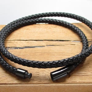 May include: A black braided leather necklace with a magnetic clasp. The necklace is coiled on a wooden surface, showcasing its texture and design. The necklace is approximately 18 inches long.