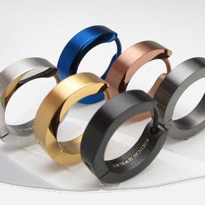 Pode incluir: Um conjunto de seis brincos de argola com vários acabamentos metálicos, incluindo prata, azul, dourado, cobre, preto e cinza chumbo. Cada brinco circular possui um fecho de dobradiça e o texto KIKUCHI JEWELRY gravado na superfície interna.
