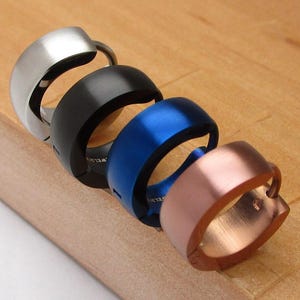 May include: Four rings in silver, black, blue, and copper are displayed on a wooden surface. The rings have a smooth, rounded design, with a slight curve. The rings are arranged diagonally, showcasing their different colours and finishes.
