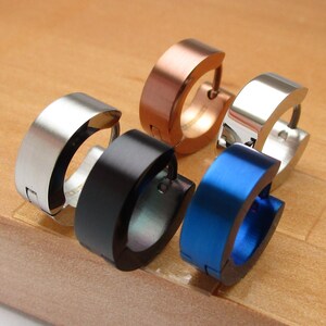 Op de afbeelding: Vijf roestvrijstalen oorringen in zilver, koper, zwart en blauw. De oorbellen liggen op een houten oppervlak. De oorbellen hebben een eenvoudig, modern ontwerp met een platte voorkant en een gebogen achterkant.