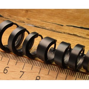 May include: A set of six black hoop earrings arranged on a wooden surface next to a ruler. The earrings are made of a dark metal and have a circular design. The earrings are of varying sizes.