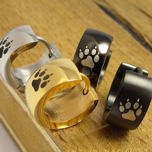 Puede incluir: Tres pendientes de aro con un diseño de huella de pata. Un pendiente es plateado, uno es dorado y uno es negro.