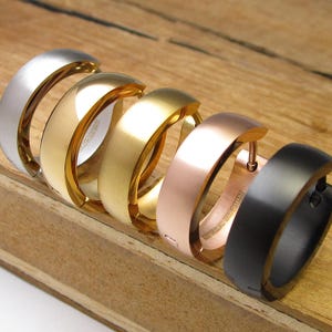Op de afbeelding: Een set van vijf oorringen gerangschikt in een rij, met een glad, afgerond ontwerp. De oorbellen zijn afgewerkt in zilver, goud, geelgoud, roségoud en matzwarte tinten, elk met een scharniersluiting en zichtbare tekst aan de binnenzijde.
