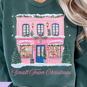 May include: Dark green sweatshirt featuring a pink shop with a blue door, Christmas decorations, and the text "Small Town Christmas." Two Christmas trees flank the shop. The image also includes the text "Bubble Bloom" and "PNG."