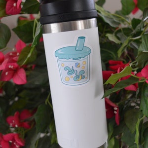 K&ouml;nnte beinhalten: Eine wei&szlig;e Edelstahl-Wasserflasche mit einem Aufkleber eines blauen und gr&uuml;nen Cartoon-Boba-Tees mit Schlangen darin. Der Aufkleber befindet sich auf der Vorderseite der Flasche.