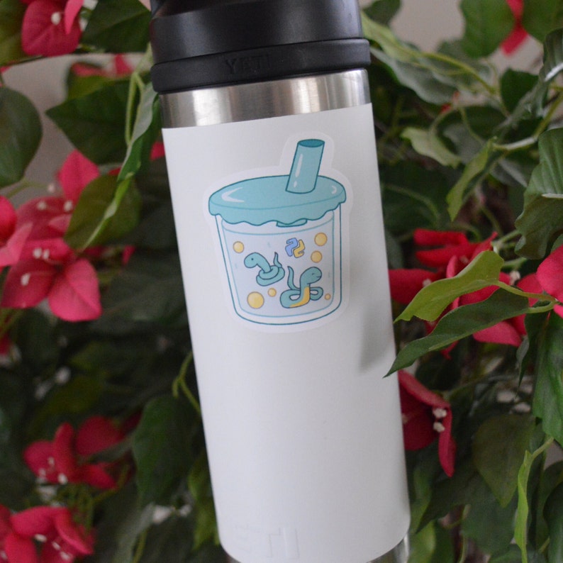 K&ouml;nnte beinhalten: Eine wei&szlig;e Edelstahl-Wasserflasche mit einem Aufkleber eines blauen und gr&uuml;nen Cartoon-Boba-Tees mit Schlangen darin. Der Aufkleber befindet sich auf der Vorderseite der Flasche.