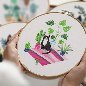 May include: Embroidered hoop art featuring a black and white cat sitting on a pink and gray striped rug surrounded by potted plants, including a cactus and a monstera plant.